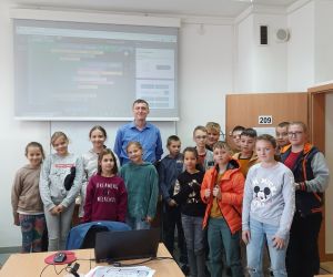 Grupa uczniów i prowadzący stoją w klasie, za nimi znajduje się ekran zawierający informacje z wykładu.