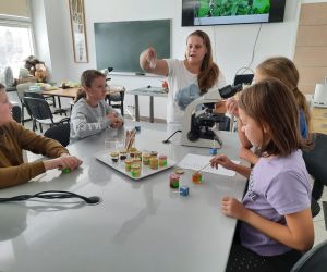 Uczniowie siedzą przy stoliku, prowadząca podaje im miody do degustacji.