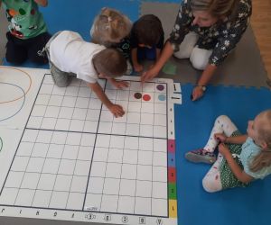 Przedszkolaki z nauczycielką układają kolorowe kropki na białej planszy w kratkę
