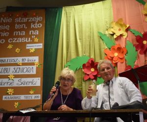 Kobieta i mężczyzna siedzą przy stole. Kobieta trzyma mikrofon.