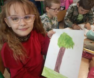 Dziewczynka w czerwonej bluzce trzyma rysunek ,na którym jest drzewo.