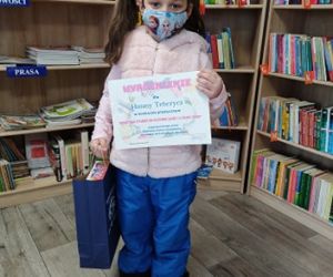 Dziewczynka na tle regałów z książkami. W ręku trzyma dyplom i nagrodę.