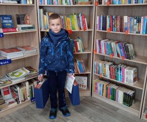 Chłopiec na tle regałów z książkami. W ręku trzyma dyplom i nagrodę.