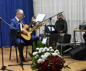 Osoba na scenie gra na gitarze, druga osoba siedzi z boku.