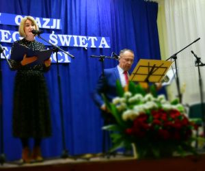 Osoba występuje na scenie. Osoby prowadzące koncert znajdują się z boku.