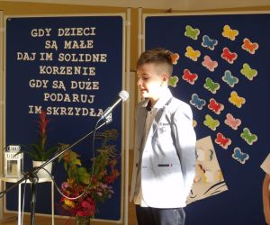 Uczeń klasy trzeciej w stroju galowym recytuje wiersz przy mikrofonie. W tle tablica dekoracyjna