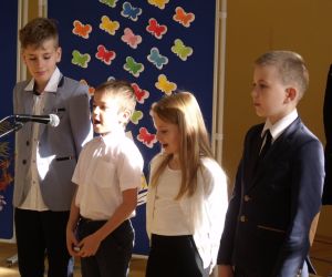 Dziewczynka z klasy drugiej i trzech chłopców z klasy trzeciej w strojach galowych śpiewają do mikrofonu