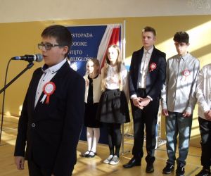 Uczeń klasy ósmej w stroju galowym przy mikrofonie. W tle pozostali uczniowie biorący udział w występie