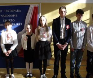 Uczniowie klasy czwartej i ósmej w strojach galowych na tle tablicy dekoracyjnej z flagą narodową