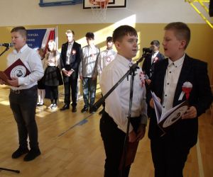 Trzech chłopców w strojach galowych przy mikrofonach śpiewa pieśń patriotyczną