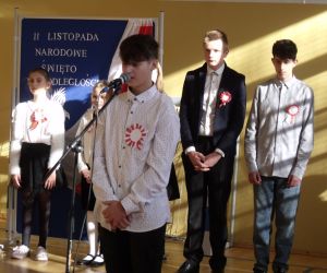 Uczeń klasy ósmej w stroju galowym przy mikrofonie. W tle pozostali występujący uczniowie i tablica dekoracyjna