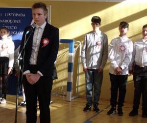 Uczeń klasy ósmej w stroju galowym przy mikrofonie. W tle pozostali występujący uczniowie i tablica dekoracyjna