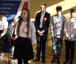 Uczennica klasy ósmej w stroju galowym przy mikrofonie. W tle pozostali występujący uczniowie i tablica dekoracyjna