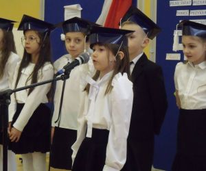 Uczennica klasy pierwszej w birecie i stroju galowym przy mikrofonie. Za nią pozostali uczniowie i uczennice