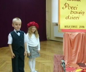 Dwoje dzieci ubranych odświętnie, pozują do zdjęcia na scenie. Tablica z napisem: Turniej Sztuki i Słowa, Poeci bawią dzieci.