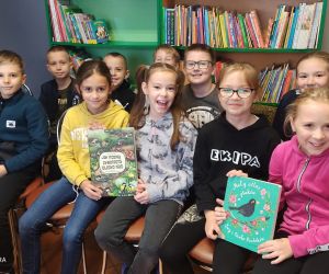 Na zdjęciu widoczne są dzieci siedzące na krzesłach, na tle regału z książkami.