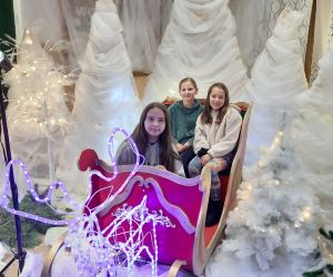 Uczennice siedzą w czerwonych saniach, za nimi stoją ośnieżone choinki