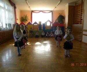 Dziewczynki stoją w dwóch, rzędach, prezentują jakiś układ taneczny, chłopcy siedzą przy oknie. 