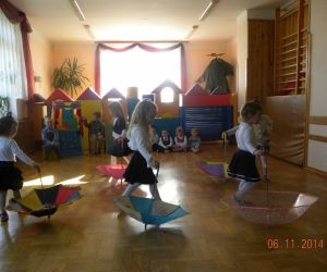 Dziewczynki prezentują układ taneczny z parasolkami, chłopcy siedzą przy oknie. 