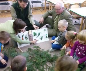 Dzieci siedzą na podłodze w kole. W środku leżą rożne gałęzie, pani nauczycielka pokazuje im coś w książce. W grupie są również dwaj leśnicy.