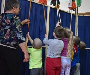 Na zdjęciu znajduje się tył parawanu koloru niebieskiego, za którym widać pięcioro dzieci trzymających kukiełki oraz jedną osobę starszą, która pomaga jednemu dziecku w animowaniu kukiełki.