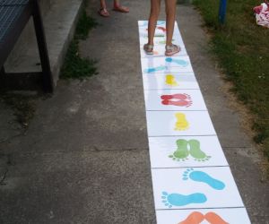 Na zdjęciu znajdują się dzieci, które uczestniczą w zabawie „wyścig stópek”. Skaczą po gigantycznej tablicy układając stopy w odpowiedniej pozycji.