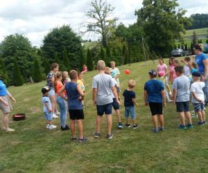 Na zdjęciu grupa dzieci stoi obok siebie tworząc okrąg. Niedaleko nich znajdują się animatorzy. Grupka dzieci podrzuca czerwony balon, który jest napełniony wewnątrz wodą.