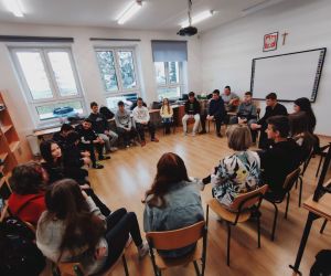 Grupa osób siedzi w kole wewnątrz budynku.