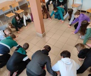 Grupa osób klęczy w kole wewnątrz budynku. Młodzież gra w grę 