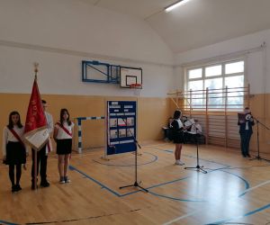 Dwoje uczniów stoi przy mikrofonach, obok stoi Poczet sztandarowy