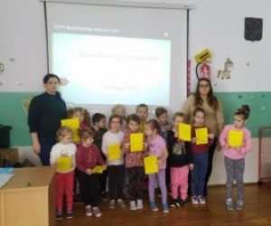 Grupa dzieci i dwie nauczycielki, stoją przy tablicy interaktywnej. Dzieci trzymają w dłoniach żółte kartki.