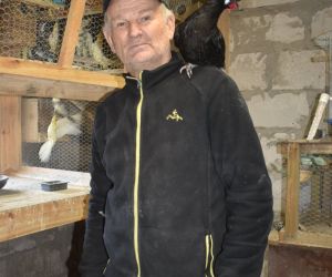Na zdjęciu znajduje się właściciel hodowli, który na ramieniu trzyma czarna kurę ozdobna, która jest oswojona. W tle klatka z papugami.