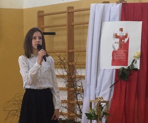 Uczennica w stroju galowym recytuje na tle dekoracji okolicznościowej