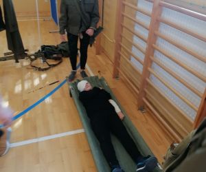 Uczennica w umundurowaniu stoi przy umundurowanym uczniu leżącym na noszach