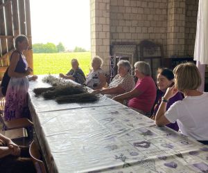 W stodole wielki stół na którym leży wysuszona lawenda, przy nim siedzą ludzie, Prowadząca spotkanie stoi,ubrana jest na fioletowo