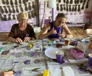 Dwie kobiety siedzą obok siebie przy stole, są zajęte przygotowywaniem pracy plastycznej, która jest tematem warsztatów