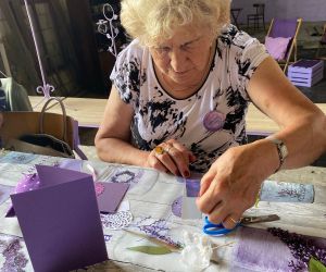 Seniorka przykleja papierowy element. Jest skupiona na wykonaniu swojej pracy.