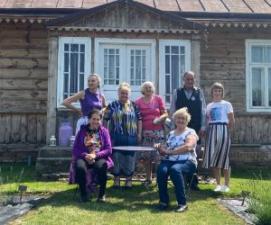 Grupa seniorów pozuje do zdjęcia wraz z prowadzącymi warsztaty. W tle drewniana chata