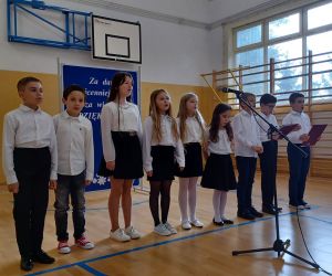 Uczniowie klasy trzeciej w strojach galowych śpiewają przy mikrofonach