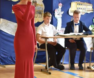 Dawna uczennica szkoły śpiewa przy mikrofonie. W tle dwóch uczniów przy stoliku.