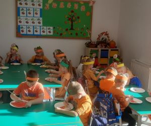 Dzieci siedzą przy stolikach w sali przedszkolnej, wykonując prace plastyczną – dynie z papierowych talerzyków. Na głowach mają własnoręcznie zrobione opaski.