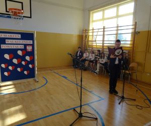 Uczeń w stroju galowym czyta przy mikrofonie; w tle tablica z dekoracją okolicznościową