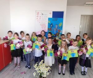 Przedszkolaki w eleganckich ubraniach trzymają w rękach bukiety kwiatów i kolorowe laurki