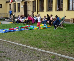 Na trawie leżą kolorowe zabawki, obok siedzą na leżakach dorosli