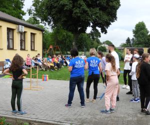 Grupa ludzi stoi obok siebie, dzieci czekają na swoją kolej w grze, dorośli pilnują zasad