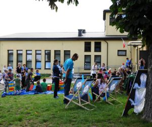 Na podwórzu jest dużo osób, dzieci się bawią a dorośli siedzą i rozmawiają ze sobą 