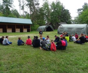 Dzieci siedzą na świeżym powietrzu, na środku prowadzący mówi do grupy.