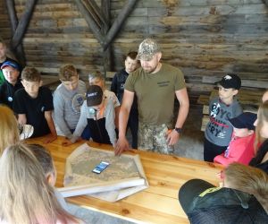 Dzieci znajdują się pod drewnianą altaną, w środku dorosły mężczyzna prezentuje mapę.