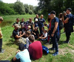 Dzieci stoją w grupie na świeżym powietrzu. W tle rzeka Bug. Dorosły mężczyzna prezentuje coś grupie.