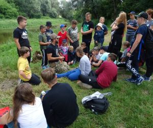 Dzieci stoją w grupie na świeżym powietrzu. W tle rzeka Bug. Dorosły mężczyzna prezentuje coś grupie.